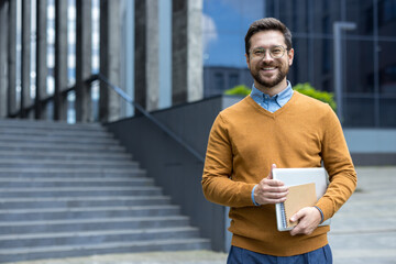 Portrait of a young businessman in a shirt, sweater and glasses standing near an office building,...