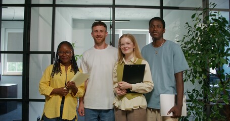 A group of young people in casual clothes are smiling at the camera while standing in the office. The startup team. Daylight