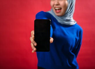 An Asian woman in a grey hijab and blue sweater holds a smartphone forward, smiling energetically...