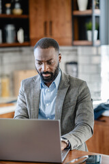 Focused African-American man working on his laptop at home, dressed in business attire. He appears...