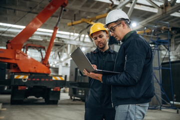 Architect is showing the plan on the laptop. Two male factory workers are in the warehouse