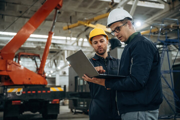 Architect is showing the plan on the laptop. Two male factory workers are in the warehouse