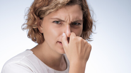 Fototapeta premium A young woman with short curly hair and a white t-shirt pinches her nose in clear discomfort, reacting to a bad smell. Her facial expression vividly conveys the unpleasantness of the odor. Ideal for