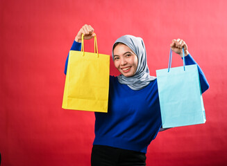 Asian woman wearing a gray hijab and blue sweater holds up yellow and blue shopping bags with a joyful expression. She stands against a red background, looking thrilled with her shopping