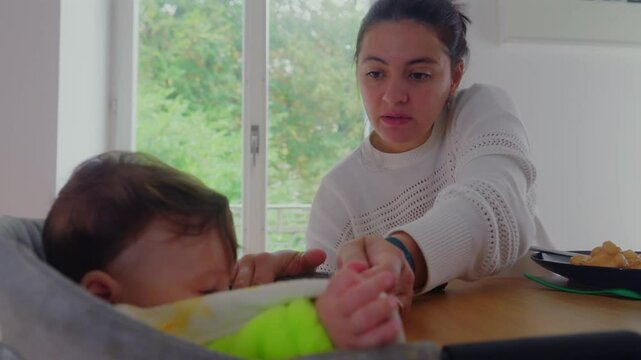 Close-up of baby eating in high chair, focus on baby&rsquo;s expression while being spoon-fed by mother, attentive look, intimate feeding moment at the dining table