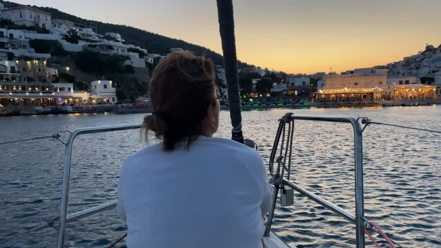 Greece,Leros Island, Panteli Bay, a woman watching the sunset from a sailboat over looking the Panteli beach and the city lights, such a peaceful time of the day.