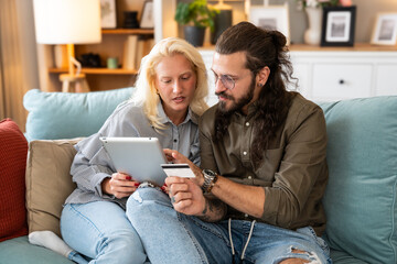 Cheerful happy young couple using digital tablet, man holding credit card, together making an...