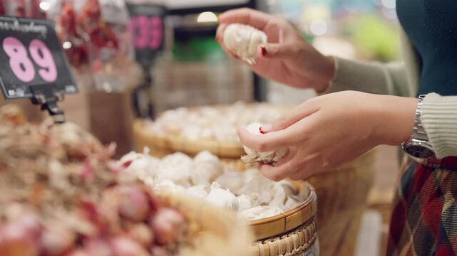 A woman is holding a bunch of garlic in her hand. She is in a market, surrounded by various fruits and vegetables. The atmosphere is lively and bustling, with people shopping