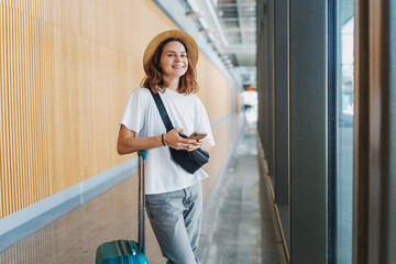 Young happy woman traveler in hat with suitcase and smartphone in hands at airport or train station waiting for travel