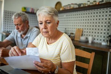 Concerned senior couple reading bills on kitchen table with laptop