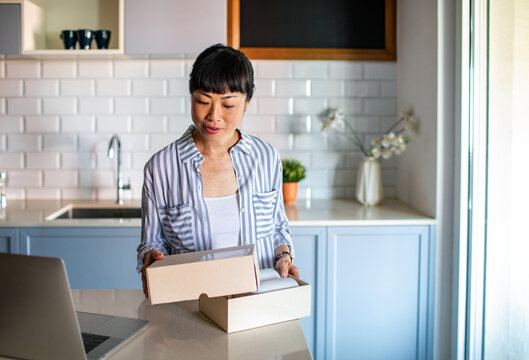 Smiling Asian woman opening package at home with laptop
