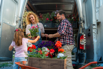 A family of three loading colorful flowers from a wheelbarrow into a delivery van for transport, highlighting their small family business.