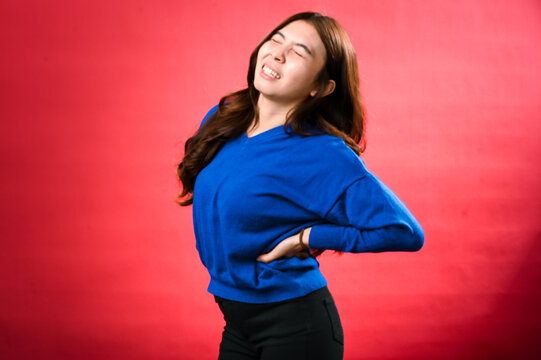 An Asian woman in a blue sweater, slightly bending backward with her hand placed on her lower back, grimacing in pain as if experiencing discomfort. The background is red