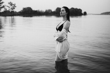 Portrait of an attractive long-haired girl walking on a beach by the water. A young woman in a swimming costume and shirt meets a summer dawn on the river. Black and white photo