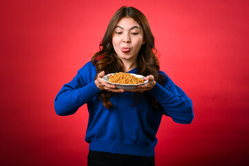 An Asian woman in a blue sweater holds a plate of noodles close to her face, licking her lips in anticipation. She stands against a red background with a playful and hungry expression