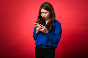 An Asian woman in a blue sweater frowns while looking at her phone, appearing confused or displeased. She stands against a bright red background, holding the phone with both hands