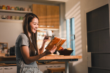young woman enjoys cozy moment reading book while sipping drink in bright kitchen. warm light...