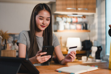 young woman is sitting at wooden table, looking at her smartphone while holding credit card. She appears focused and engaged, surrounded by cozy home setting with various items in background