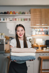 young woman smiles while holding stack of neatly folded towels in cozy kitchen setting. warm ambiance and her cheerful expression create welcoming atmosphere