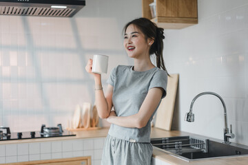 woman in gray outfit stands in modern kitchen, holding white mug and smiling. bright space features sleek appliances and wooden accents, creating cheerful and inviting atmosphere