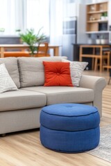 Fototapeta premium A stylish living room features a light gray couch adorned with orange and patterned pillows. A round blue ottoman sits nearby, adding a pop of color to the warm, inviting space filled with natural lig
