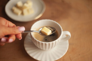 Butter coffee or bulletproof coffee, a high-calorie, high-fat drink free from carbohydrates to start the day; a woman's hand adding a spoonful of butter to a cup of coffee