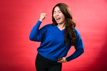 Fototapeta premium An Asian woman in a blue sweater and black pants is standing against a red background, smiling confidently while raising her fist in a gesture of strength and victory, her other hand on her hip