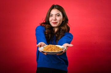 An Asian woman wearing a blue sweater is enthusiastically holding out a plate of noodles toward the...