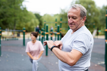 Ssenior man in sportswear looks at fitness watch or at pedometer while exercising in city park
