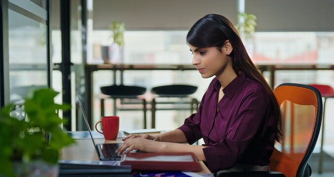 Indian young adult gen z busy woman lady use laptop look screen typing check email indoor job office place teen girl female expert staff sitting chair do online work task search project data ideas	