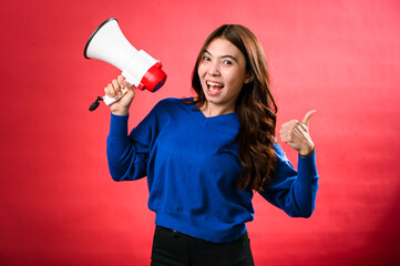 An Asian woman wearing a blue sweater is holding a red and white megaphone while speaking into it. She appears to be shouting or making an announcement. The background is solid red