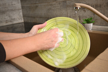 Hands washing a green plate with a yellow sponge under running water in a kitchen sink