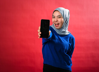 An Asian woman in a grey hijab and blue sweater holds a smartphone forward, smiling energetically...