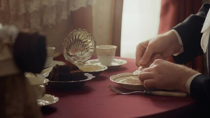 Closeup of aristocratic man in formal attire taking butter from vintage shell shape butter dish with knife and putting it on piece of bread on porcelain plate while having lunch in wealthy atmosphere