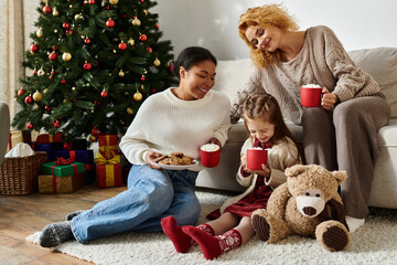 A lesbian couple shares festive treats and warm drinks with their daughter by the tree.