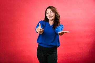 An Asian woman standing against a red background, wearing a blue sweater and black pants. She is giving a thumbs-up with one hand while extending the other hand forward with her palm open