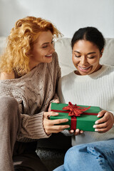 Two women express happiness while exchanging a wrapped gift in their cozy living space.