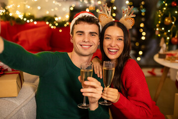 Happy European couple cheering with champagne glasses and making selfie, sitting in living room interior with Christmas decorations and garlands