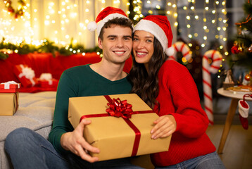 Romantic European spouses holding wrapped gift box and smiling at camera, celebrating Christmas holidays at home