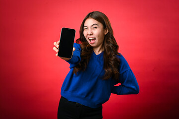 Naklejka premium An Asian woman wearing a blue sweater is smiling excitedly while holding a smartphone forward in her hand, displaying its screen to the viewer. The background is a bold, solid red