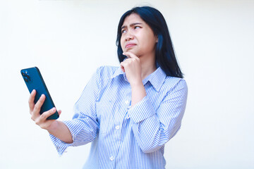 thoughtful asian woman using smartphone with hands on chin thinking problem wearing blue striped casual shirt standing over isolated white background, look away