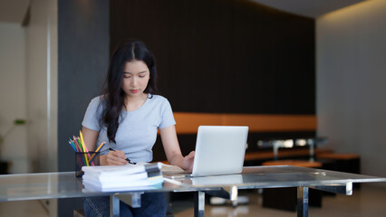 Asian teenage student woman reading a book to taking notes for assignment homework while watching lecture video online lesson on laptop to studying and learning about education knowledge in cafe