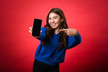 An Asian woman in a blue sweater is smiling and pointing at a smartphone she's holding forward, displaying its black screen. The background is a bold red, creating a striking contrast