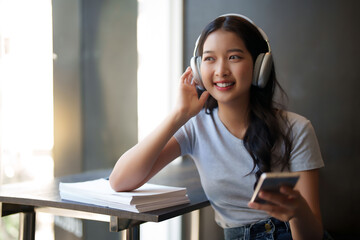 Asian teenage student woman wearing headphone to listening music and surfing social media on smartphone to relaxation with happiness after studying education knowledge for preparing test exam in cafe