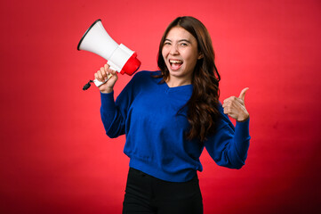 An Asian woman wearing a blue sweater is holding a red and white megaphone while speaking into it. She appears to be shouting or making an announcement. The background is solid red