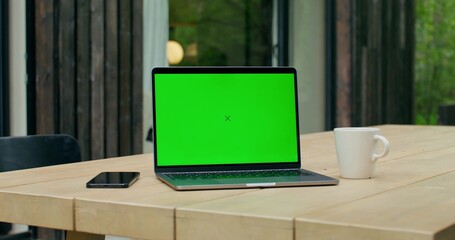 An open laptop with a green screen is on a table on the terrace of a stylish cottage.