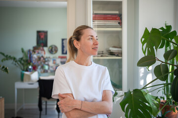 Engrossed in dreamful thoughts middle aged woman calm looks to window stands at home spending...