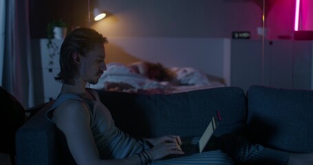 A man uses a laptop sitting on the couch at night. A woman is sleeping in a bed. Dark room, home interior