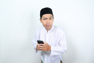 closeup of muslim asian man holding mobile phone looking at camera with confused expression isolated on white background