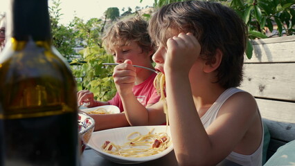 Two children enjoying an outdoor meal, focusing on the boy stirring spaghetti on his plate with a fork. Casual family dining setting with greenery in the background